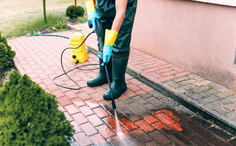 Man cleaning red, conrete pavement block using high pressure water cleaner. Paving cleaning concept. Man wearing waders, rubber boots, gloves and waterproof trousers doing spring jobs in the garden